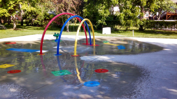 The Splash Pad at Eise Park (three rings of decreasing size, from red to blue to yellow respectively amongst various multicolored water spouts in concrete) lie in a pool of water. In the background, a field, trees, and a playground are in the sunlight.